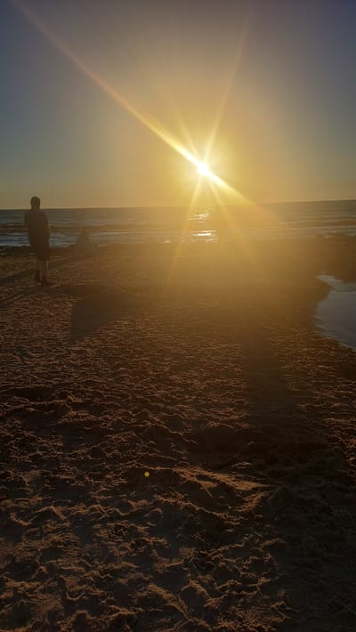 A wide shot captures the sunset at Vääna-Jõesuu beach in Estonia. The sun, a bright starburst, dominates the upper right quadrant, casting a warm golden glow across the scene. The sky transitions from a clear blue near the left horizon to a hazy yellow and orange near the sun. The sea stretches across the middle ground, its surface reflecting the sunlight in shimmering patterns. On the shore, the sand is a rich brown, textured with ripples and shadows. In the foreground, a person stands facing the ocean, their silhouette dark against the bright sky. Further back, another figure sits on the sand, also facing the water. A small, dark pool of water is visible on the sand to the right, reflecting the sky. The overall impression is one of tranquility and natural beauty.