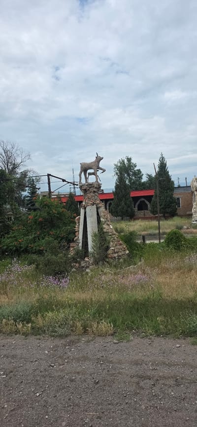 In Sasyk-Bulak, Kyrgyzstan, a stone and concrete sculpture depicting a deer and a fawn stands atop a weathered, triangular pedestal constructed from rocks and concrete. The sculpture is situated in a grassy, overgrown area with scattered wildflowers. Behind it, a building with a red arched roofline and dark window openings is visible, partially obscured by trees. The sky above is overcast. A gravel path runs in the foreground of the image.
