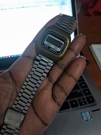 A person holds a retro digital watch with a stainless steel band in their hand. The watch face is rectangular and features an LCD display. The word "Quartz" is visible on the face. The background shows a laptop, with part of the keyboard and a sticker visible. The hand holding the watch is in focus, while the laptop in the background appears slightly blurred.
