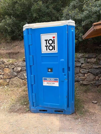 A blue plastic portable toilet, manufactured by TOI TOI, is situated outdoors in Montanejos, Spain. The toilet sits on a dirt and gravel ground surface, adjacent to a stone wall. The environment suggests a rural or semi-rural location, evidenced by the presence of uncultivated ground and the stone structure. A patch of vegetation, likely bushes or trees, is visible to the left of the frame.