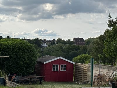 The image shows a red wooden shed with white-framed windows in a yard. A wooden picnic table and bench are to the left of the shed. In the background, there are trees, buildings, and a church with a steeple. The sky is cloudy. The scene is located in Stolberg, Germany.