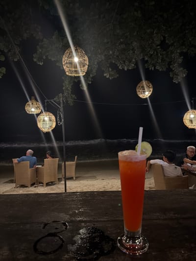 The image captures a nighttime scene at a beachfront establishment in Ao Phai, Thailand. In the immediate foreground, a tall glass containing an orange-red frosted beverage, garnished with a white straw and a lime wedge, sits on a dark, wet surface, possibly a wooden bar top, which exhibits a visible numeral "3" and scattered moisture droplets.
The midground reveals a sandy beach area furnished with wicker tables and chairs. Several individuals are seated, facing the ocean. On the left, two individuals with their backs to the camera are seated at a table. On the right, two males are seated, one younger individual in a white polo shirt with his back mostly to the camera, and an older male with white hair and glasses partially visible.
The background features the dark expanse of the ocean with distinct white-capped waves breaking on the shore. Overhead, multiple spherical light fixtures, appearing to be crafted from woven rattan or bamboo, are suspended from dark, leafy tree branches and a metal pole structure. These fixtures emit bright white light, creating prominent light rays that cut through the dark night sky. The overall scene depicts a casual, illuminated outdoor dining or lounge area beside the sea.