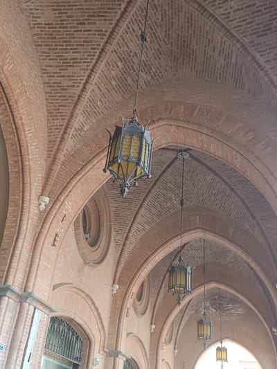 The image shows the interior of an arched brick walkway in Casalmaggiore, Italy. Three ornate, wrought-iron lanterns with yellow glass panels hang from the ceiling, illuminating the space. The walkway is constructed with reddish-brown bricks arranged in a vaulted ceiling pattern. Circular decorative windows with metal grilles are visible on the right side of the walkway. The lower portions of the walkway reveal arched doorways with decorative metalwork. There are no people or animals visible in the image.