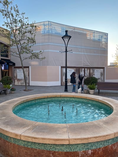 The scene is an outdoor commercial plaza located in Marratxí, Spain, specifically an area resembling the Mallorca Fashion Outlet. A circular fountain, with a concrete lip and a base clad in brown and teal mosaic tiles, occupies the lower foreground, containing still, clear blue water with multiple metal nozzles.
Beyond the fountain, a paved walkway extends. A black, ornate street lamppost stands prominently in the midground. To the left, a deciduous tree with nascent green leaves and dark spherical fruits is situated near a building exhibiting a "Carhartt" branded awning. To the right of the lamppost, two individuals are observed with a small white dog. One person, attired in a dark jacket and blue jeans, maintains control of the dog via a leash. The second person, wearing a grey jacket and jeans, is bending forward and downward, appearing to interact with the dog or an object on the ground. Adjacent to this interaction are several potted plants, including a fern and shrubs. A brown slatted wooden bench is positioned further to the right.
The background is dominated by a multi-story commercial building under renovation or construction. Its upper sections are enveloped in light-colored protective sheeting and scaffolding, with glimpses of underlying glass structures. Lower sections of the building feature light brown panels covering storefronts, adorned with "Pepe Jeans" promotional posters. The sky above is a clear, light blue, indicative of daylight.