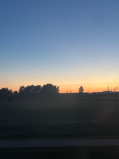 The image depicts a rural landscape at dusk. The sky transitions from a clear blue at the top to a warm orange glow near the horizon. Silhouetted against the sky are a line of trees and a distant industrial area with smokestacks emitting faint plumes of smoke. In the foreground, a dark, horizontal strip, possibly a road or a field boundary, is visible. The overall scene is peaceful and serene, characteristic of a twilight in a Latvian countryside. The specific location in Jaunolaine, Latvia, is suggested by the presence of agricultural fields and typical Latvian industrial structures seen in the distance. There are no people or animals visible in the image.