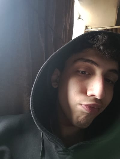 A young individual, appearing male, is depicted in a close-up indoor portrait. The subject, positioned centrally, has dark, curly hair and dark eyes, and is wearing a dark hooded sweatshirt with the hood up, framing the face. The individual's expression involves a direct gaze towards the camera with lips slightly pursed. Illumination appears to originate from the upper right, highlighting the right side of the face. The background consists of a dark, vertically oriented curtain or fabric on the left, and a lighter-toned ceiling or upper wall area on the right, which includes a glimpse of a structural element or fixture. The image provides no discernible features to confirm a specific location within Mankat al-Hatab, Syria.