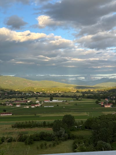 The image displays a wide, horizontally oriented landscape, which appears to be rotated 90 degrees clockwise from its natural upright position. The left portion of the frame is dominated by a dramatic sky, featuring a dynamic blend of dark, heavy grey clouds and brightly illuminated white and golden-tinged clouds. Patches of clear blue sky are visible, breaking through the cloud cover, suggesting either a late afternoon/early evening light or a change in weather conditions.

The right side of the image unveils a vast, verdant valley bordered by layers of rolling, tree-covered mountains and hills in the mid-distance, extending across the horizon with a slightly hazy appearance. The valley floor is intricately patterned with numerous rectangular plots of green agricultural fields and pastures, indicating active cultivation. Scattered throughout this lush landscape are small clusters of houses, mostly featuring red or brown roofs, characteristic of a rural settlement or village. Dense foliage, including individual trees and wooded areas, is abundant, particularly along the hillsides and bordering the fields.

No people or discernible text are visible in the scene. The overall impression conveyed is one of natural beauty and rural tranquility, likely capturing the expansive landscape around Miločani, Montenegro, bathed in a soft, atmospheric light.
