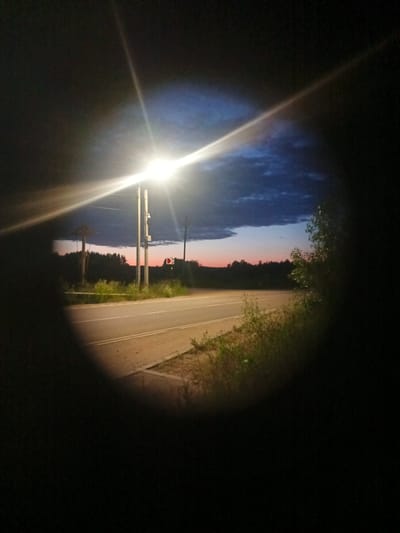 The image, captured at dusk in Voskresenskoye, Russia, depicts a street scene framed by a dark, vignetted border. A bright streetlamp illuminates the foreground, casting strong beams of light. To the left, a wooden structure with a pointed roof stands near a road. A traffic sign indicating a direction is visible on the right side of the road. The road itself is paved and appears to be a two-lane highway, bordered by grassy areas and trees in the distance. The sky shows a gradient of colors, transitioning from a deep blue with clouds at the top to lighter hues of pink and orange near the horizon. The overall impression is one of a rural or semi-rural setting at the beginning of the night.