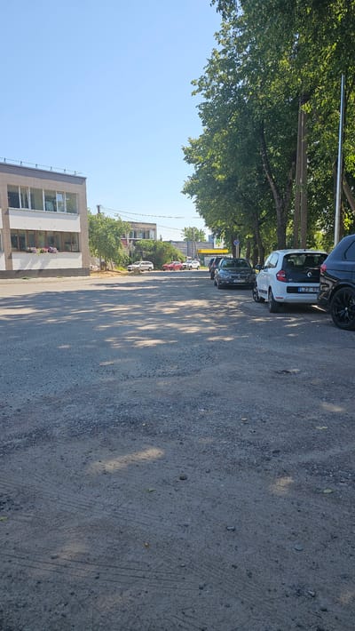 The image depicts a sunny day in a parking lot in Nemėžis, Lithuania. A modern building with a light-colored facade and multiple windows is visible on the left side of the frame. Adjacent to the building is a line of trees casting shadows on the asphalt and gravel parking surface. Several cars are parked along the right side of the lot, including a white Renault Twingo with a license plate "LZZ 671", a dark BMW, and a black SUV. In the background, a yellow awning of a commercial establishment can be seen, and more trees line the street. The ground of the parking lot is predominantly gravel with some patches of asphalt, showing tire tracks and scattered small stones. The sky is clear and blue.