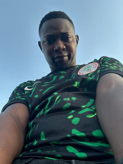 A low-angle, waist-up, outdoor selfie is captured against a clear blue sky. The subject is a Black man wearing a black and green patterned Nigeria national football team jersey. He has short, dark hair and a light beard. His left arm and shoulder are visible in the foreground, and he is looking down at the camera. The image is sharp and well-lit, with no apparent distortions.