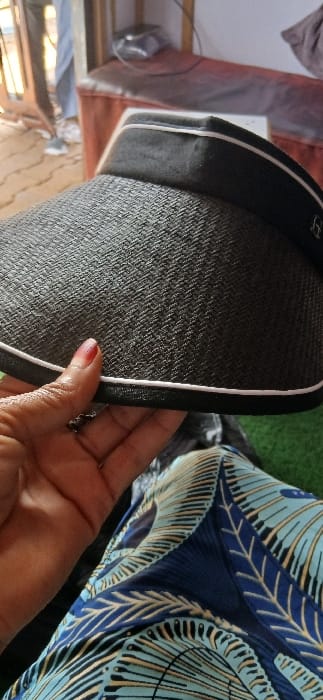The image captures an indoor scene in Sabon Gari South, Nigeria, looking downwards. In the foreground, a person's right hand, adorned with red nail polish and a ring on the ring finger, holds a black woven visor with a white trim along its edge and a plain black headband. Below the hand, a section of a leg or lap is visible, covered by fabric featuring a light blue, dark blue, and cream patterned design. The immediate surface beneath the person is green, consistent with artificial turf. The background is blurred. To the left, partial lower legs and feet wearing light-colored athletic shoes are visible through a wooden-framed opening, standing on an outdoor paved surface with a light-colored brick pattern. To the right, a piece of furniture upholstered in reddish-brown fabric is present, with an indistinct object and cabling resting on its surface.