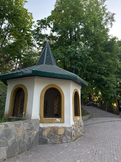 The image depicts a small, white structure with a pointed, green-tiled roof, possibly a chapel or shelter, located outdoors. The structure features arched windows with gold trim and metal grilles. The base of the structure is made of stacked stones. The setting appears to be a park or wooded area, with tall green trees visible in the background. A paved walkway with steps is partially visible to the right of the structure. The image likely depicts a location in Krasny Klyuch, Russia, as requested.
