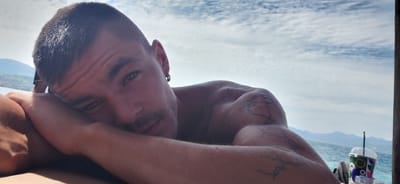 This is a first-person, close-up, selfie-style photograph of a man resting his head on his arm. He has short dark hair, stubble, and a ring earring in his left ear. He is shirtless and appears to be wet, with water droplets visible on his skin and shoulder. A tattoo is visible on his upper left arm. The background consists of a bright, cloudy sky over a body of water, with mountains visible in the distance. The man is positioned such that his face is on the left side of the frame, looking towards the viewer. A colorful drink with a straw is visible in the lower right corner, suggesting he is outdoors and possibly on a boat or near a waterfront establishment. The scene is set against the backdrop of the Municipal Unit of Aegio, Greece, likely a coastal area given the water and mountainous terrain.