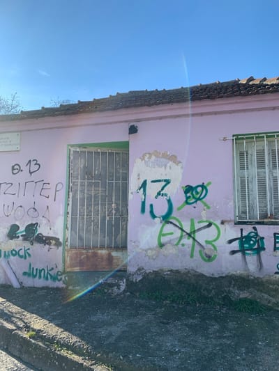 The image displays a side view of a weathered building with a light pink facade and a reddish-brown tiled roof, set against a clear blue sky. The wall shows significant paint peeling and is covered in various graffiti.
Two windows are visible on the building's facade. The upper window has light-colored slatted shutters, while both the upper and a lower, smaller window are protected by metal security grates, some of which appear rusty. The building's base features a green trim.
Prominent green graffiti is scrawled across the pink wall, including a large, stylized number "13" near the center, and other abstract shapes and letters. Towards the bottom of the wall, more graffiti is visible: "θ. 13", "ΤΖΙΤΤΕΡ" (likely Greek for "Tzitter"), "WODA", and "rock Junkies". A small, rectangular plaque is affixed to the lower left wall, below "θ. 13", with faded Greek text that is difficult to decipher fully, possibly mentioning an organization or individual.
The scene is brightly lit, indicative of daytime, with a distinct rainbow-colored lens flare arcing across the image, suggesting direct sunlight hitting the camera. The ground adjacent to the building appears to be a concrete path or dirt. Based on the location context, this scene is in Kastraki, Greece.