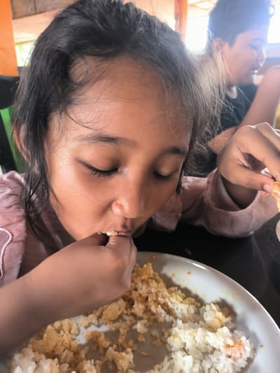 A young girl is prominently featured in this close-up shot, deeply engrossed in eating a meal of rice with her hands. She is positioned towards the left of the frame, bending over her plate with her eyes closed or cast downwards, fully focused on scooping food to her mouth. Her dark hair is somewhat dishevelled around her face, and she wears a light pink, long-sleeved shirt with noticeable ruffles or texture.
The plate before her contains a mixture of white rice and rice mixed with a reddish-orange seasoning or sauce, indicating a flavorful dish. She uses her right hand to bring the rice to her mouth, with some grains visible around her lips. Another person, appearing to be a young individual, is visible in the blurry background to the right, also seated, suggesting a shared meal in a casual setting.
The environment appears to be indoors, possibly a dining area or a restaurant, with some wooden structures visible in the upper background, characteristic of local architecture. The lighting suggests it is daytime, with natural light illuminating the scene. The overall atmosphere conveys a simple, everyday moment of sustenance. There is no visible text in the image. The location context provided is Meurah, Indonesia, which aligns with the cultural practice of eating with hands.
