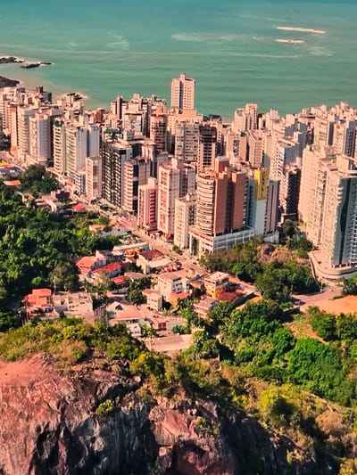This is an aerial view of a coastal city, likely Natal, Brazil, with a dense urban landscape situated between a rugged cliffside and a body of water. The urban area is characterized by numerous tall buildings, primarily residential high-rises, clustered together. These buildings vary in height, color, and architectural style, with many featuring balconies and modern designs. Below the high-rises, there is a lower-density residential area with smaller houses, many with red-tiled roofs, interspersed with lush green trees and vegetation. The terrain slopes upwards from the sea, with the city built on the slopes. A significant rocky cliff face dominates the foreground, covered in sparse vegetation. The water appears calm and is a greenish-blue color, with some small waves and rocky outcrops visible near the shore. The overall impression is of a densely populated city that has grown organically along a scenic coastline, with a clear demarcation between the built environment and the natural landscape.