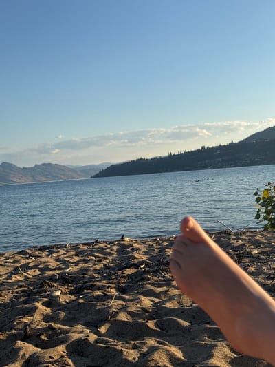 The image depicts a serene beach scene at Okanagan Lake in Kelowna, Canada. In the foreground, a bare foot is positioned on the sandy shore, casting a shadow on the textured sand. The sand is littered with small pieces of driftwood and debris. The middle ground is occupied by the calm, blue waters of Okanagan Lake, with a flock of birds visible in the distance. The background features a lush, tree-covered hillside that slopes down to the water, with mountains forming a majestic backdrop against a clear, pale blue sky. A few wispy clouds are scattered across the horizon. The lighting suggests it is either early morning or late afternoon, with the sun casting long shadows.