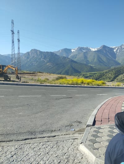 A wide-angle, eye-level, outdoor shot captures a scenic view of the city of Dardar, Uzbekistan, on a clear, sunny day. The foreground is dominated by a grey asphalt road with white lane markings, curving gently to the right. A paved sidewalk with a red brick and decorative grey tile pattern borders the road on the right. In the middle ground, a bright yellow excavator is parked on a patch of dirt and vegetation. Beyond the road and excavator, a vibrant field of yellow wildflowers stretches across the landscape. The background features a majestic mountain range with green slopes and snow-capped peaks under a clear blue sky. Two tall, industrial power pylons stand prominently on the left side of the frame, with power lines extending across the scene. The overall impression is one of a natural landscape meeting industrial infrastructure.