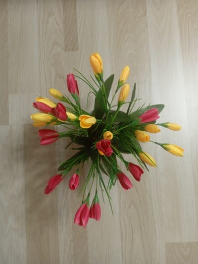 A top-down, medium shot shows a bouquet of artificial tulips, a mix of red and yellow, arranged on a light-colored wooden floor. The bouquet is centered in the frame, with the tulips fanning out from a central point. Some of the tulips are fully closed buds, while others are partially opened. Green artificial leaves and stems are interspersed among the flowers. The wooden floor has a visible grain pattern. There are no people or animals present in the image. The scene is indoors. The location in Unkurgan, Uzbekistan, is not discernible from the image itself.