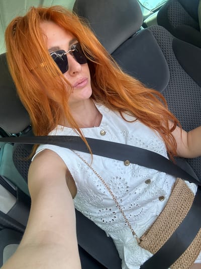 This media file is a selfie of a woman with long, auburn hair, wearing sunglasses and a white eyelet top. She is seated in a car, with a seatbelt across her body. A woven straw bag with a chain strap is visible over her shoulder. The background shows car seats. The lighting suggests it is daytime. The image is oriented sideways. The location context provided is Novoselje, Montenegro. There is no visible text in the image.
