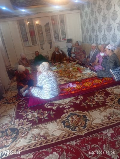 In Чоң-Гара, Kyrgyzstan, a group of women and children are gathered for a meal or social event on a large, intricately patterned carpet. The room features a large, ornate cabinet with glass doors displaying various items, suggesting a domestic setting. The women are seated on the floor, some wearing traditional headscarves. Several platters of food are arranged on the carpet, indicating a meal is in progress. The overall atmosphere appears relaxed and familial.