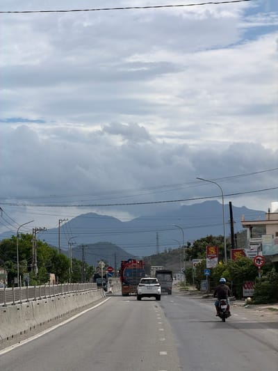 The image depicts a daytime scene on a road in Ba Ngoi Ward, Vietnam, with mountains in the background under a cloudy sky. The perspective is looking down a straight section of asphalt road, which has white dashed lines indicating lanes and a solid white line on the right shoulder. A concrete median barrier with regularly spaced metal posts separates the road from the right-hand side.

On the road, several vehicles are visible. In the foreground, a white pickup truck, possibly a Toyota Hilux, has a large, blue and red cylindrical tank or container secured on its bed. Behind it, a darker, possibly black, truck can be seen. Further down the road, a person wearing a dark shirt and a light-colored helmet rides a motorcycle, moving away from the viewer. The motorcycle's license plate "63B3 033.39" is partially visible, while the white pickup truck's plate starts with "51C-".

The right side of the road is lined with trees and power poles carrying numerous electrical wires. Buildings are partially visible behind the trees and power lines, suggesting commercial or residential areas. Several signs are present: a red circular sign indicating "No U-turn," and a blue triangular sign depicting two figures, likely a pedestrian or children crossing warning. Advertisements and business signs include "QUÁN CƠM THANH HÒA" (Thanh Hoa Rice Restaurant), "TẠP HÓA THANH HÒA" (Thanh Hoa Grocery Store), and red banners with the text "SÀI GÒN" (Saigon), likely for a beverage brand.

The weather appears overcast, with large, diffuse clouds dominating the sky, but no immediate signs of rain. The mountains in the distance are hazy, possibly due to atmospheric conditions or distance. Streetlights are mounted on curved poles above the road. The overall impression is a functional, busy thoroughfare connecting an urban or semi-urban area with a more natural, mountainous landscape.