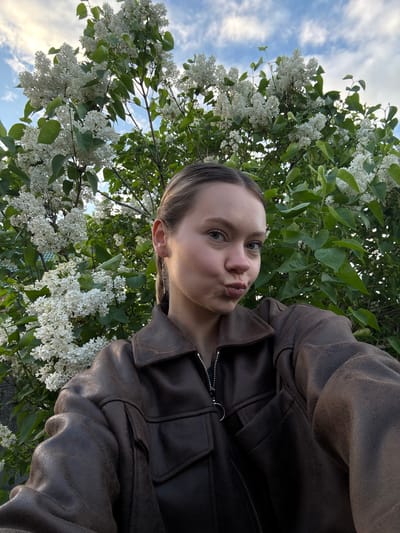 A young woman poses for a selfie outdoors, possibly in Severnyi, Russia, though this is not definitively identifiable from the image alone. She wears a brown leather jacket and has her hair pulled back. Her lips are pursed in a kissing face expression. Behind her are branches of a flowering bush with white blossoms, possibly lilac. Above and behind the bush, the sky is visible with scattered clouds.