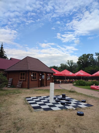 The image depicts an outdoor recreational area with a wooden building, a checkerboard game, and an outdoor dining area. The wooden building, possibly a cafe or restaurant, has a brown, shingled roof and is constructed from logs. Two ladders lean against its side. In front of the building is a large checkerboard laid out on the ground, with several black checker pieces scattered on it. To the right of the checkerboard, there's a red beanbag chair with the Coca-Cola logo. Beyond that, a paved path leads to an outdoor seating area covered by large red umbrellas, also bearing the Coca-Cola logo. Several tables and chairs are set up under the umbrellas, and some patrons are seated. The background is filled with lush green trees. The sky is blue with scattered white clouds. The location is identified as Shcherbani, Ukraine.