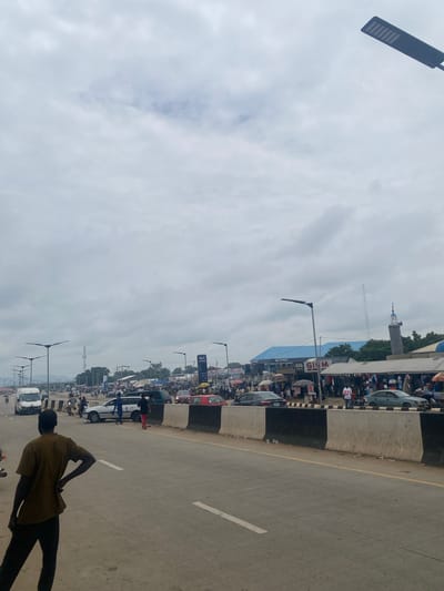 A wide shot captures a bustling urban scene along a highway in Pasere, Nigeria, under a heavily overcast sky. The foreground is dominated by a paved road with white lane markings and a concrete barrier separating the traffic lanes from the roadside market. A man in a brown shirt and dark pants stands with his back to the camera, facing the road.
On the right side of the road, stalls are set up with awnings, displaying various goods, likely clothing and accessories. Numerous people are browsing and shopping. Beyond the market, a tall, slender structure resembling a mosque minaret with a blue-capped top is visible. A large blue building with white lettering that reads "GIG LOGISTICS" and "ENUGU" is situated behind the market stalls. Further down the road, vehicles are moving, including cars, vans, and motorcycles. Traffic appears moderate to heavy.
Streetlights with modern, angular designs are mounted on tall poles along the highway, illuminating the area. In the distance, trees and a faint mountain range can be seen. The overall atmosphere is that of a busy commercial area adjacent to a major thoroughfare.