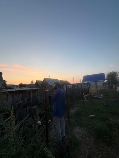 The image depicts a rural yard in the village of Kuseevo, Russia, during a sunset. A person is standing in the center of the frame, facing away from the camera, wearing a welding mask and a blue shirt, and appears to be welding a metal fence post. The person is standing on a red platform. In the background, there are several rustic wooden houses with corrugated metal roofs and a newer house with a blue metal roof. The yard is overgrown with grass and weeds, and there is a weathered wooden fence on the left side of the image. The sky is clear with faint orange and pink hues from the setting sun.