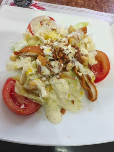 The image shows a close-up view of a Caesar salad on a white plate. The salad contains lettuce, croutons, crispy chicken strips, bacon bits, and a creamy dressing. Sliced tomatoes are placed on the sides of the plate. The salad is presented on a table, with a blurred background that suggests an indoor setting, possibly a restaurant. The overall impression is that of a prepared meal. There is no discernible location in the city of Polígono Industrial De Quart De Poblet, Spain, visible in the image.