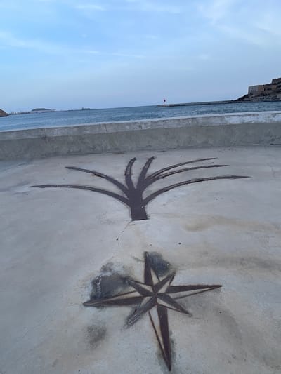 The image shows a concrete ground with two metallic engravings. In the foreground, a compass rose design is etched into the concrete. Behind it, a stylized tree-like shape branches out with each branch pointing towards a different city, with the city name and distance inscribed on it. The names visible include "ISTANBUL 2.537 Nm", "HONG KONG 9.171 Nm", "LIVORNO 937 Nm", "MADRID 322 Nm", "CARTAGENA DE INDIAS 7.330 Km", "NUEVA YORK 6.443 Km", and "ROTTERDAM 1.509 Nm". In the background, the sea is visible beyond a concrete barrier, with a lighthouse and a fortified building on a hill to the right, indicative of the city of Cartagena, Spain. The sky is cloudy with a light blue hue. There are no people visible in the image.