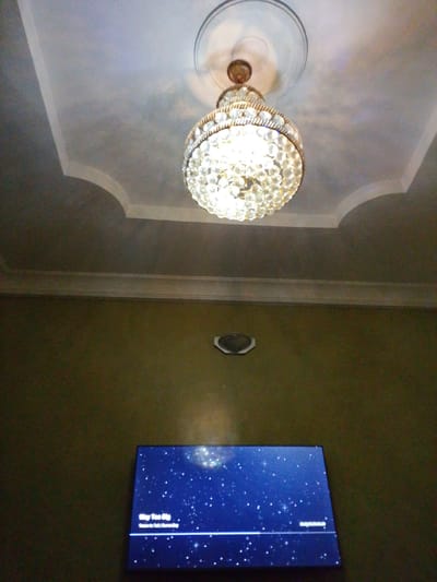 The image depicts an indoor scene featuring a ceiling fan with a decorative, crystal chandelier hanging below it. The ceiling has decorative molding. Below the ceiling, a flat-screen television is mounted on a dark green wall. The television displays a starry night sky with some text, including "Way Too Big" and "Take It All / Heaven Sky." A small, circular vent or speaker is also visible on the wall above the television. The image appears to be taken from a low angle, looking upwards. The overall impression is of a room with a somewhat ornate light fixture and modern entertainment technology.