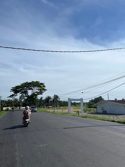 This media file depicts a scene on a road in Bukitrata, Indonesia, under a bright, sunny sky with scattered clouds, suggesting a warm daytime. The road appears to be paved asphalt, with a slight curve visible.

The primary subjects are a person riding a motorcycle and a parked car. The motorcyclist, wearing a helmet and a light-colored outfit, is moving along the road. A dark-colored car is stationary on the right side of the road. In the background, a building with a white facade and a distinctive yellow "G" logo is visible, along with some traffic cones. Beyond the building, palm trees and other vegetation are abundant, indicative of a tropical or subtropical environment. A large tree with a full canopy stands prominently in the middle ground. Power lines are a significant visual element, running across the upper portion of the frame. A white archway structure is also present in the background.

The activity appears to be everyday transportation and commerce, with the motorcyclist traveling and the building potentially being a commercial establishment. The overall mood is one of a typical day in a populated, developing area. No discernible text, other than the logo on the building, is clearly legible.
