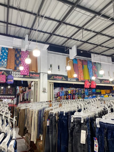 This image depicts the interior of a clothing store, possibly a small boutique or market stall, located in Lamteumen Barat, Indonesia. The scene is oriented vertically, with the ceiling structures at the top and the floor at the bottom of the frame, suggesting the photo was taken with the camera tilted.
The store is filled with various items of clothing, primarily pants and fabrics, displayed on racks and shelves. On the left side, stacks of colorful fabrics with floral and geometric patterns are visible, illuminated by bright overhead lights. These fabrics appear to be folded and arranged, possibly for sale as material or as part of a garment display.
To the right, numerous pairs of pants, predominantly jeans and casual trousers, hang on white hangers arranged on metal racks that extend deep into the store. The pants are displayed in various colors, including dark denim, black, grey, and lighter shades like beige and khaki. Some pants have visible price tags or branding.
Further into the store, more clothing items and accessories are visible, including what appear to be backpacks or bags in bright colors, and other garments displayed on shelves. The overall impression is one of a busy retail environment stocked with a wide array of apparel.
Prominently displayed on the left wall are large signs with the text "MW" and "SERBA 35.000" and "SERBA 75.000", indicating "All for 35,000" and "All for 75,000" in Indonesian currency (likely Rupiah), suggesting promotional pricing. The lighting suggests it is daytime, but the interior nature of the store makes it difficult to determine the exact time or external weather conditions. The setting appears to be a covered market or a shop with a corrugated metal roof structure. There are no people clearly visible in the frame, but the presence of merchandise and signage indicates an active retail space.