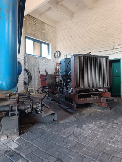 In Vuhlehirsk, Ukraine, inside a sparsely lit industrial building with white brick walls and a concrete floor, a large, rusted metal radiator unit sits mounted on a red metal base. The radiator is situated in the center of the frame, with its vertical metal fins dominating its front surface. To the left of the radiator, a large blue pipe extends upwards and out of frame, and to its right, a closed green door is visible. In front of the radiator, on its red base, is a stencil with Cyrillic text and numbers that read "ИНВ. № 30466 НБ-10". The overall environment appears to be a workshop or storage area, with various mechanical components and equipment visible in the background. The lighting is dim and casts long shadows, suggesting either early morning or late afternoon.