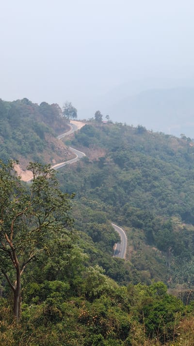 An aerial view shows a winding mountain road in Lâm Sơn, Vietnam. The road has sharp turns and is surrounded by dense green trees. The sky is hazy. A tree with green leaves is visible in the foreground.