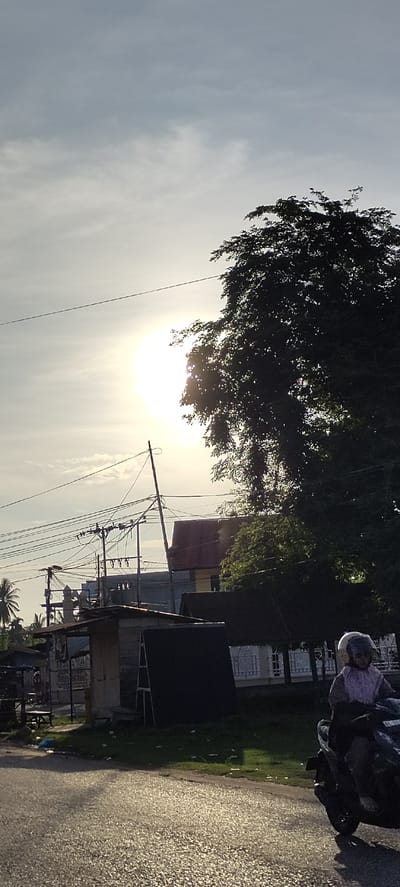 The image captures a street scene in Sumbokrayek, Indonesia, bathed in the warm glow of a setting or rising sun. The sky is a pale gray with diffused sunlight, indicating a hazy or overcast condition. A large, dark silhouette of a tree dominates the right side of the frame, its leaves catching some of the light. In the foreground, a dark asphalt road reflects the sunlight, creating a textured surface.

On the right side of the road, a person wearing a helmet and a light-colored garment is riding a black motorcycle. The person appears to be a passenger, judging by their posture and the presence of a driver (though the driver is largely obscured).

To the left of the road, there are several structures. Closest to the road is a small, open-sided shelter with a dark roof and possibly a wooden or metal frame. Behind this, a building with a dark roof and yellow walls is visible, with a red-tiled roof on its upper section. Further left, there are more buildings, including a structure with a blue roof, and palm trees. A complex network of electrical wires and poles stretches across the middle ground, suggesting the area is serviced by electricity. Scattered debris, including plastic bags and other refuse, is visible on the grassy area bordering the road. The overall impression is a modest, functional urban or semi-urban environment in Indonesia.