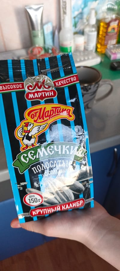 A person's hand is holding a package of "Martin" brand sunflower seeds. The package is blue and black with blue stripes and features a cartoon chicken holding a sunflower seed. Text on the package indicates "sememchki" (sunflower seeds), "polosatyye s solyu" (striped with salt), and a net weight of 150g. The background is blurred and appears to be a kitchen or bathroom counter with various bottles and a mug. The image was taken in Tikhvin, Russia.