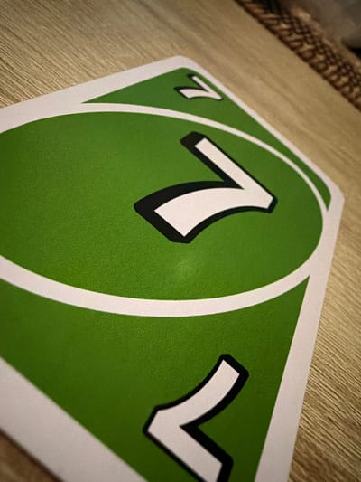 The image shows a close-up, angled view of two green UNO cards on a wooden surface. The cards have a large white "7" printed on them with a black outline. The lighting suggests the scene is indoors, possibly a game night or casual setting. There is no indication of location in Castillo Bajo-conejito, Spain, or any people present. The environment is limited to the cards and the wooden table.