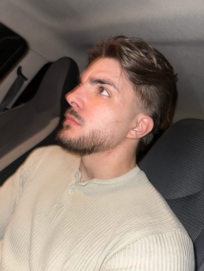 A young man with dark hair and a beard is lying down in what appears to be the back seat of a car. He is wearing a beige ribbed shirt with buttons. The seat is upholstered in a dark gray fabric. The location of the image is not discernible from the given visual information.