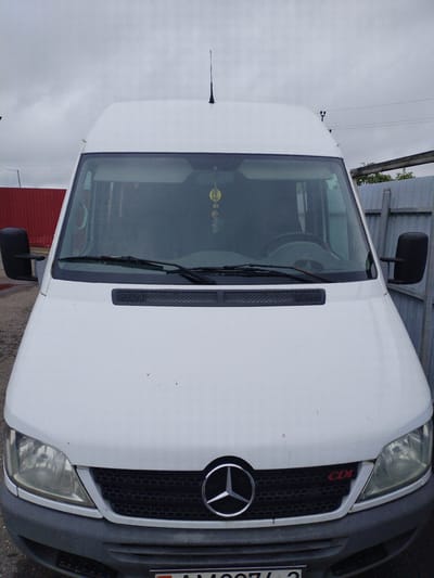 The image shows the front of a white Mercedes-Benz Sprinter van parked outdoors. The van is facing the camera, and its headlights, grille, and hood are clearly visible. A black side mirror is attached to the driver's side of the vehicle. In the background, a grey metal fence and a red fence are visible, suggesting an urban or industrial setting. The sky is overcast, and the lighting is diffused. The ground appears to be paved, with some scattered debris. A small red "CDI" badge is visible on the right side of the grille. There are no people visible in the image. The location is identified as Вароніцкі сельскі Савет, Belarus.