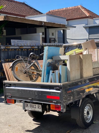 The image shows a flatbed Daihatsu Gran Max 1.5 pickup truck loaded with various items, including lumber, blue panels, a bicycle, and what appear to be furniture parts. The truck is parked on an asphalt surface, likely a street, in front of a residential area with houses featuring terracotta tiled roofs and fenced yards. The license plate of the truck is visible and reads "DK 8841 AS". Based on the license plate prefix "DK", the image is located in Bali, Indonesia. The presence of houses with distinct architectural styles, the style of the gate, and the license plate format suggest the location is within the city of Denpasar Barat, Indonesia. The scene is brightly lit by sunlight, indicating it is daytime. There are no people visible in the image.