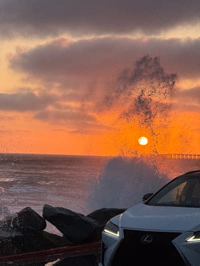 A large wave crashes against rocks in front of a white Lexus SUV parked on a street. The sun is setting over the Pacific Ocean, casting an orange glow on the clouds and water. The sky is filled with dramatic, dark clouds, and the setting sun is partially obscured by the spray from the wave, creating a halo effect. A distant pier is visible on the horizon. The scene is captured in a vertical, close-up shot, with the car occupying the foreground and the ocean and sky filling the background. The location appears to be Oceanside, California, given the presence of a pier and the ocean.