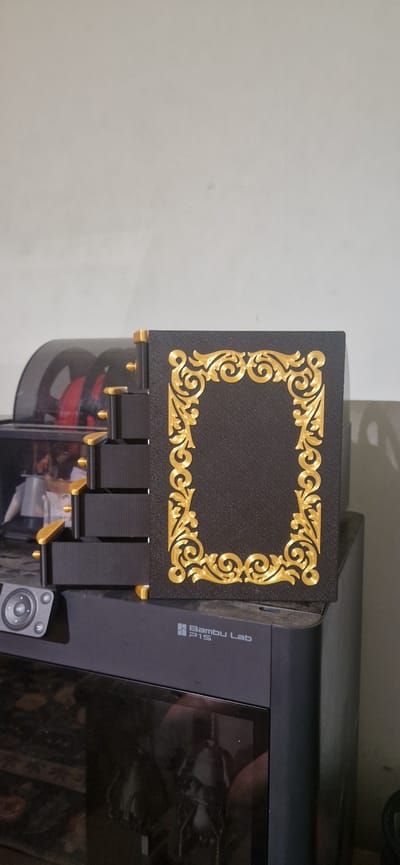 A black, rectangular box with ornate gold trim resembling a book is placed on top of a black 3D printer. The box has a textured surface, and the gold trim features intricate swirling patterns and floral motifs. To the left of the box, a series of black drawers with gold accents are visible, stacked vertically. These drawers appear to be part of a larger unit or the 3D printer itself. In the background, a large black reel with red filament is partially visible, suggesting it is a spool for the 3D printer. The scene is set against a plain, light-colored wall. The 3D printer has the "Bambu Lab P1S" logo printed on its front.
