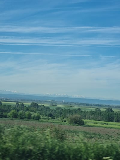 The image presents a landscape scene likely taken from a moving vehicle, with the view skewed due to the angle.
The sky occupies a significant portion of the left side, displaying a light blue color interspersed with streaks of contrails or high cirrus clouds.
The right side features a panoramic view of a terrain that transitions from flat lands to mountains in the distance. It is likely the land surrounding the city of Kozar Belene, Bulgaria. Along the lower margin, a dense green area, potentially a field of crops or grassland, is visible.
Further away, a row of trees delineates a boundary, and beyond that lies a mixture of green fields and possibly some built structures, indicating a settled area. In the far distance, mountains can be seen on the horizon, their peaks possibly snow-capped. The atmospheric conditions appear hazy or slightly overcast, muting the colors and blurring distant details.