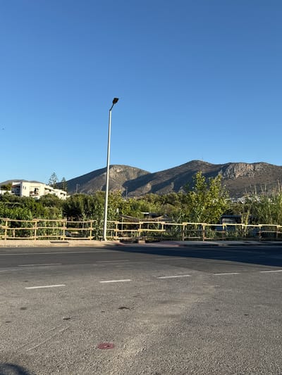 A road view captures a clear blue sky over a distant mountainous landscape, likely within the Chersonisos Municipal Unit, Greece. In the foreground, asphalt pavement with white lane markings runs horizontally. A simple wooden barrier, possibly part of a bridge or walkway, separates the road from vegetation and a few buildings. A tall, slender streetlamp stands vertically on the right side of the road, its light fixture at the top. The background features rolling hills and mountains, with some sparsely populated structures visible on the slopes. Lush green trees and tall grasses grow behind the wooden barrier. The scene is well-lit, suggesting it was taken during daylight hours.