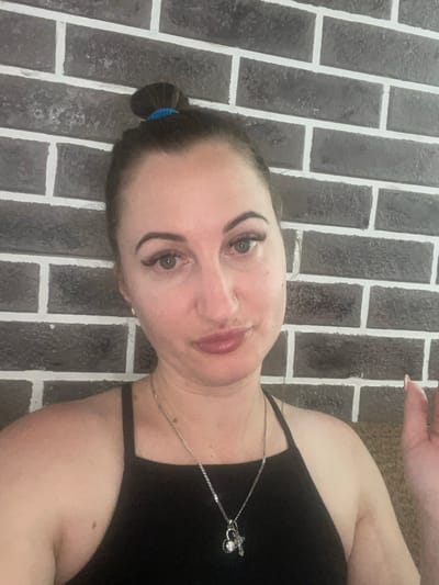 A medium shot captures a woman from the shoulders up against a dark gray brick wall with white mortar. She has her hair pulled up into a bun at the top of her head, secured with a blue hair tie. Her face is turned slightly towards the camera, with her eyes looking forward. She is wearing a black, sleeveless top with thin straps, and a silver necklace with a pendant is visible. The lighting appears to be artificial, casting a soft glow on her face and creating subtle shadows. There are no identifiable landmarks or contextual clues within the image to suggest a specific location in Zarichne, Ukraine.