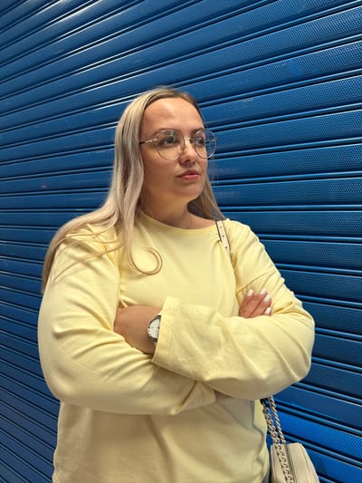 A woman with blonde hair and glasses stands with her arms crossed in front of a blue metal grate. She is wearing a yellow long-sleeved shirt and a white watch on her left wrist. A light-colored purse with a chain strap is visible on her right side. The background is a solid blue, textured surface composed of horizontal slats with small perforations. The lighting appears to be artificial and comes from the front, casting soft shadows. The scene is captured from a medium-full shot. This image was taken in Ermakovo, Russia.