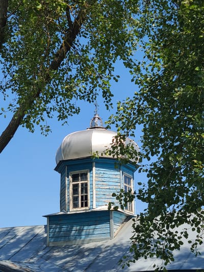 A domed bell tower with a cross on top rises above a blue wooden structure, which is part of a church in Mala Pereshchepyna, Ukraine. The bell tower is topped with a silver, bulbous dome, and a cross is visible at its apex. Below the dome, a square section of the tower is painted blue with peeling paint, and features a window with a white frame. This blue structure sits on top of the main body of the church, which has a metal roof. The church is partially obscured by the branches and leaves of a large tree in the foreground. The sky is clear and blue.